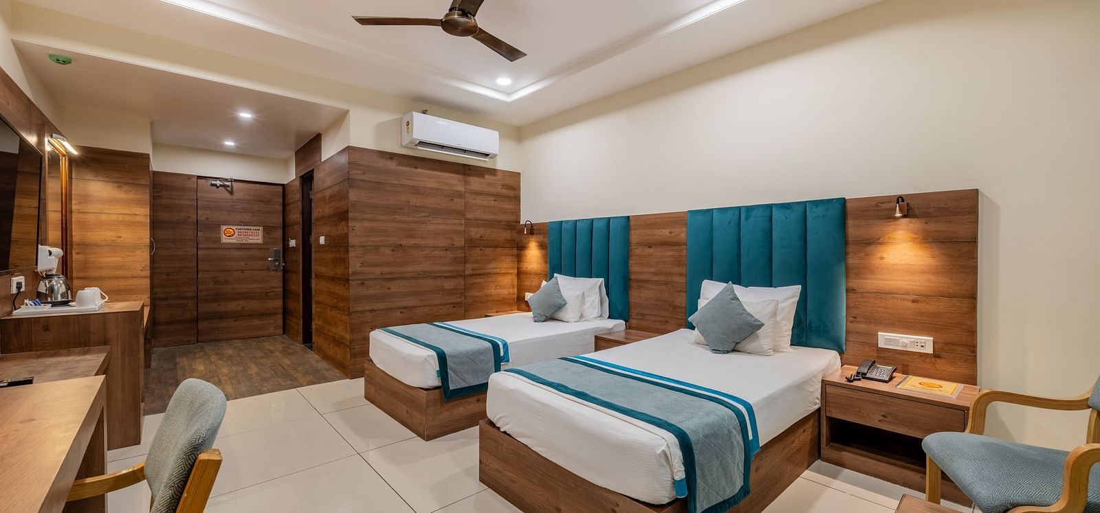 View of a King twin bed room with polished flooring, teal padded headboards, seating area, work station, ceiling fan, and ambient ceiling lights at Hotel Highway King, Bilaspur.