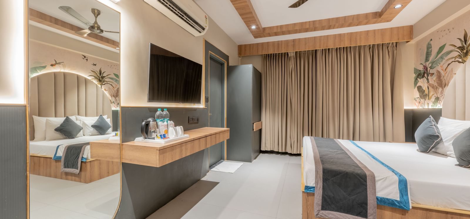 A room with a king size bed, ceiling to floor curtain, wall-mounted TV, wooden desk, and a mirror at Hotel Highway King, Jaipur.