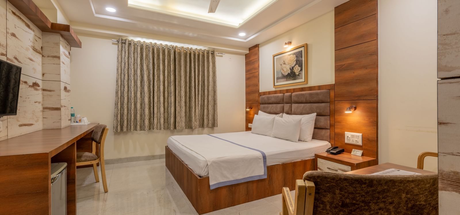 A room featuring a bed with white and blue linen, a wooden desk and chair, and a wall art at Hotel Highway King, Jaipur.