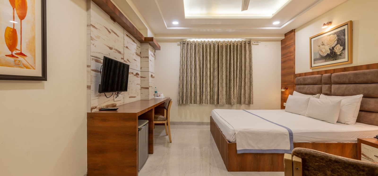 A room featuring a king size bed, wall arts, a wooden desk with a chair, and a mounted television, illuminated by recessed lighting at Hotel Highway King, Jaipur.