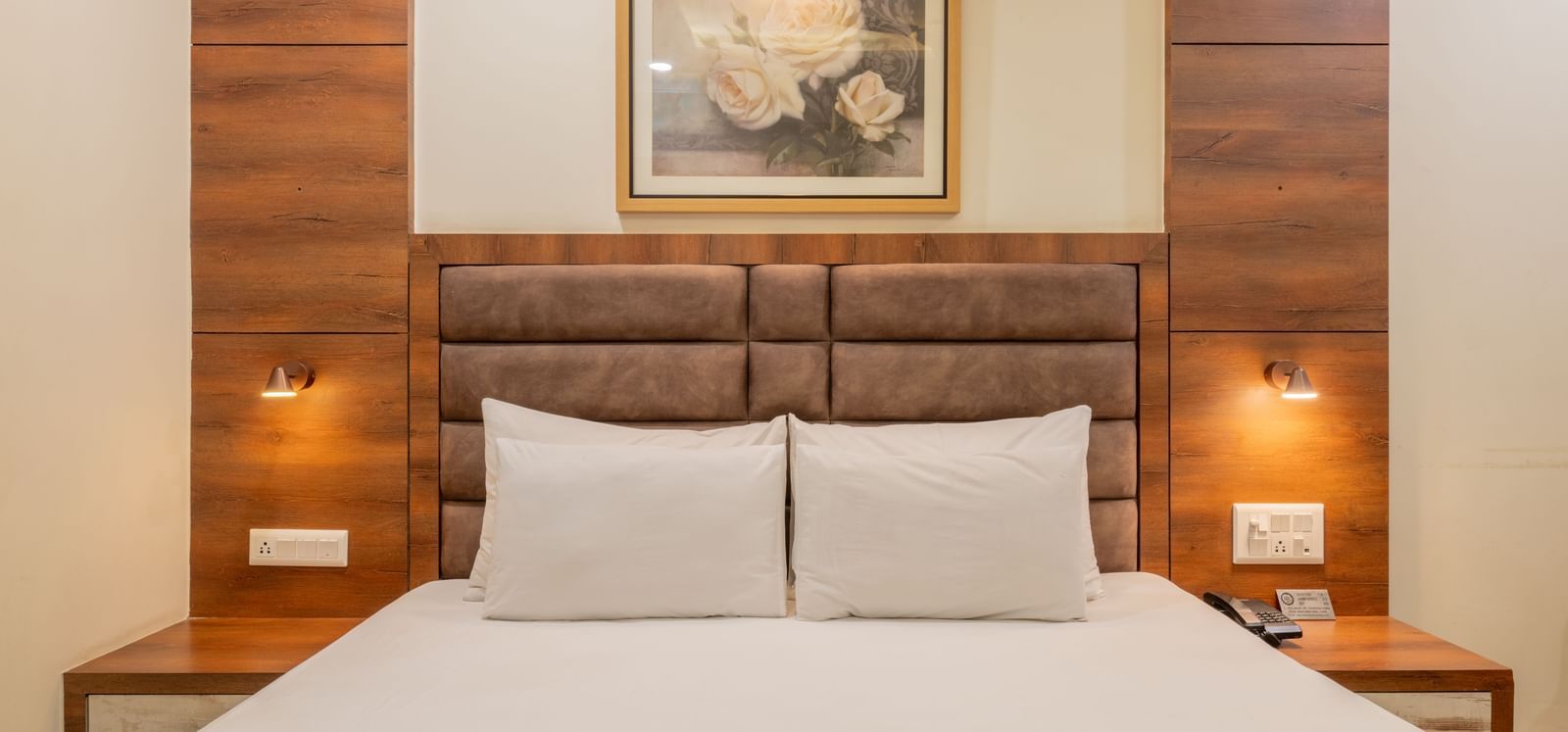 A close-up view of king size bed with padded headboard, two wall lamps, and framed artwork above at Hotel Highway King, Jaipur.