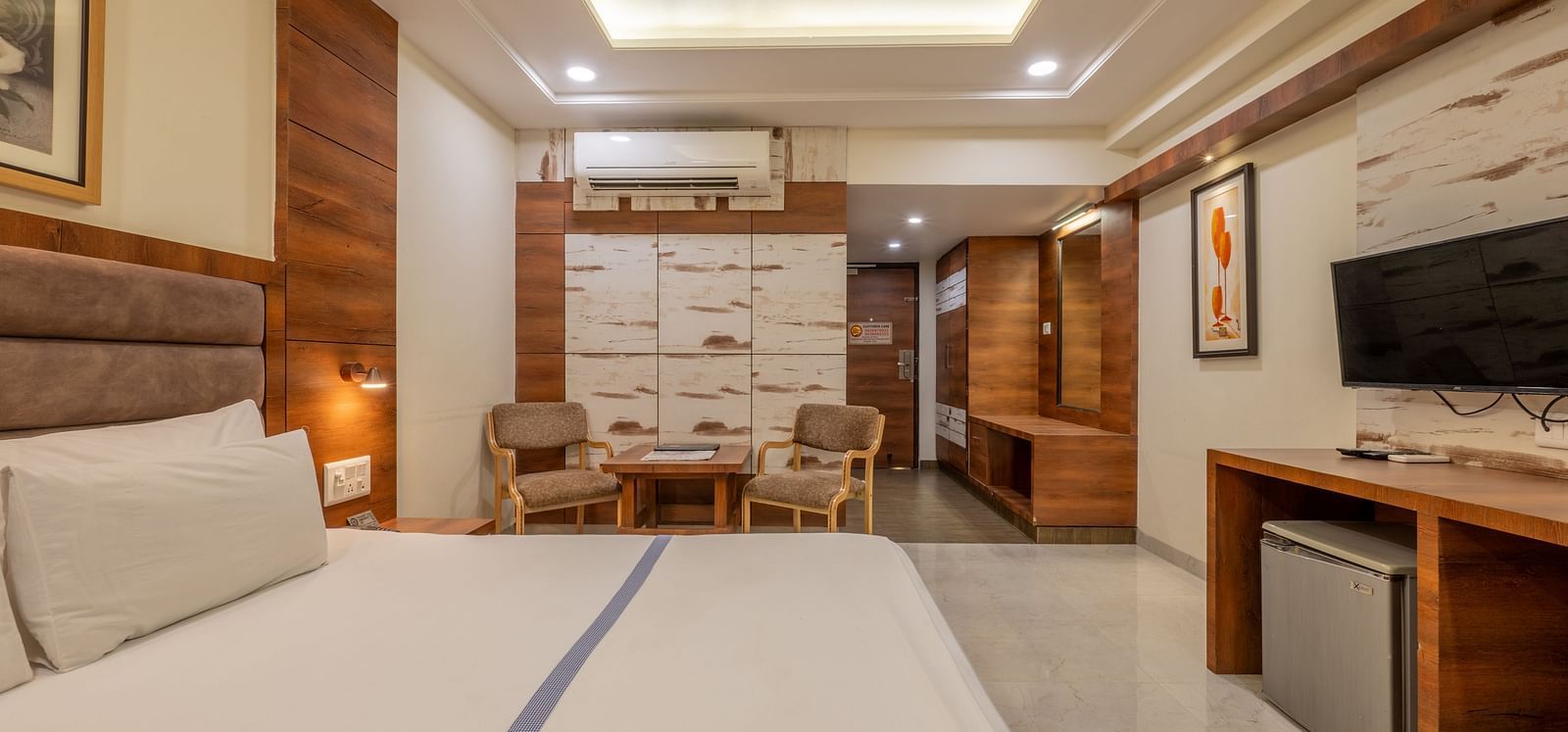 A wide view of a modern room, featuring a king size bed, a TV area, chairs and a table space, and wooden furnishings at Hotel Highway King, Jaipur.