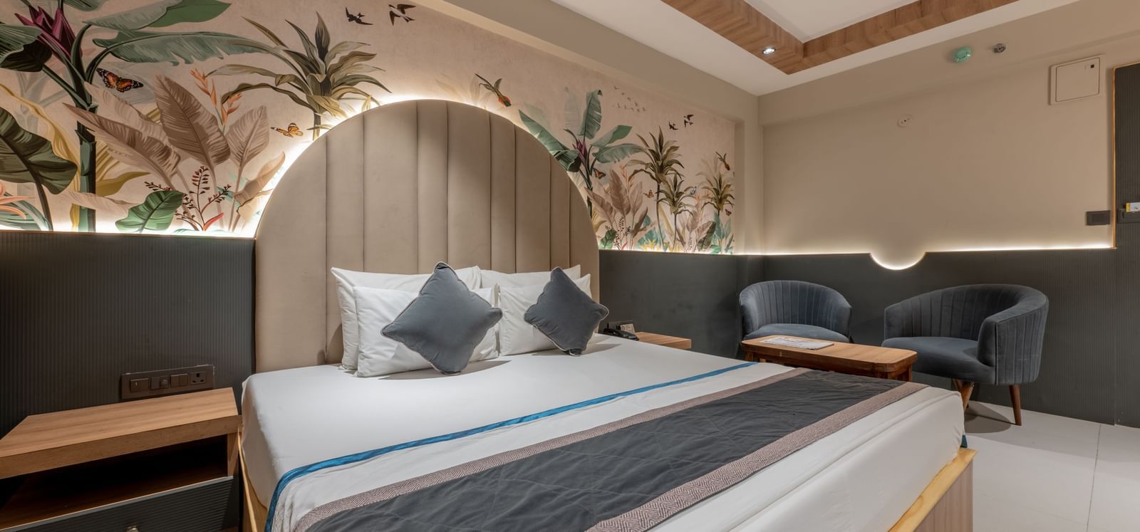 A room with king size bed, ceiling fan, floral mural wall, and grey chairs at Hotel Highway King, Jaipur.