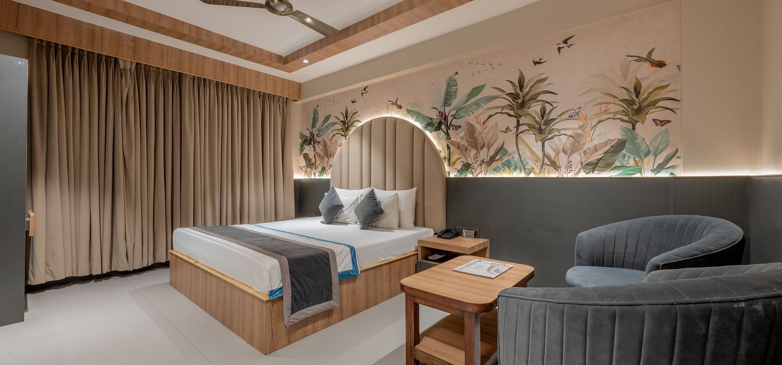 A wide view of the room with a king size bed, brown curtains, grey chairs, a wooden table and a flowery mural on the wall at Hotel Highway King, Jaipur.