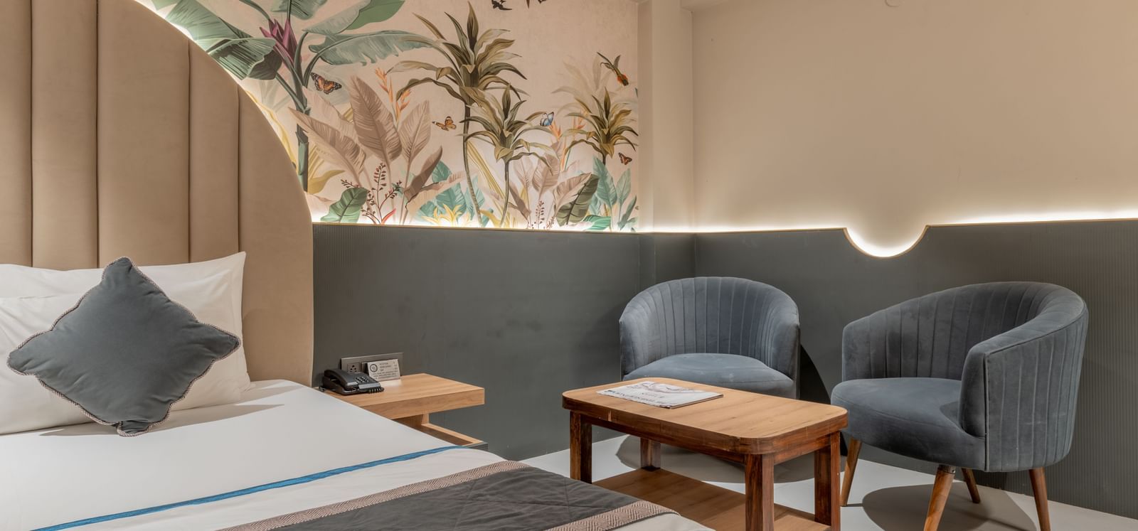 A seating area with grey chairs and wooden table beside bed and floral wall design at Hotel Highway King, Jaipur.