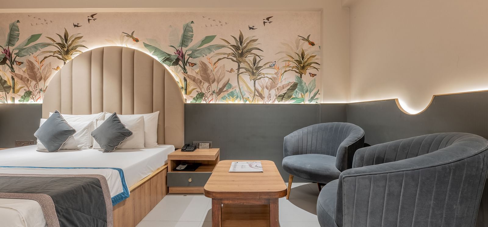 A partial view of a king size bed featuring grey accents, grey armchairs, a wooden table and plant-themed wall mural in a well-lit room at Hotel Highway King, Jaipur.