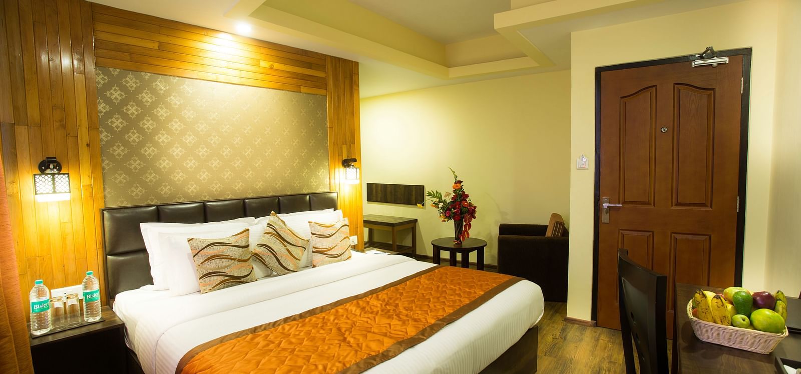 A room at New Orchid Lords Inn Gangtok contains a bed with pillows, bedside tables with lamps and water bottles, a desk with a chair and fruit bowl, and a door.