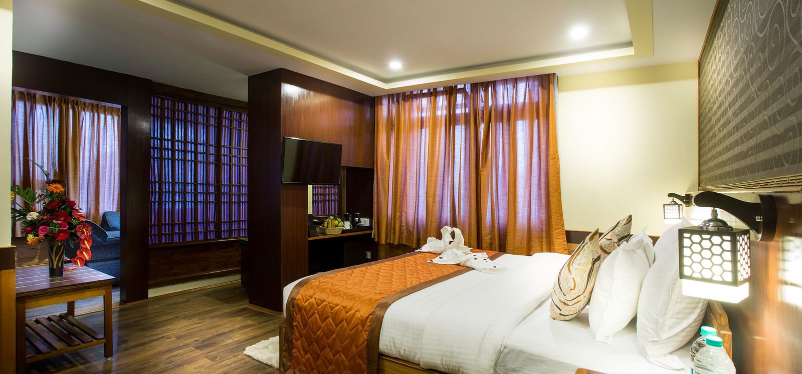 A room at New Orchid Lords Inn Gangtok contains a bed with pillows and folded towels, bedside tables with lamps and water, curtains, a table with flowers, and a television.