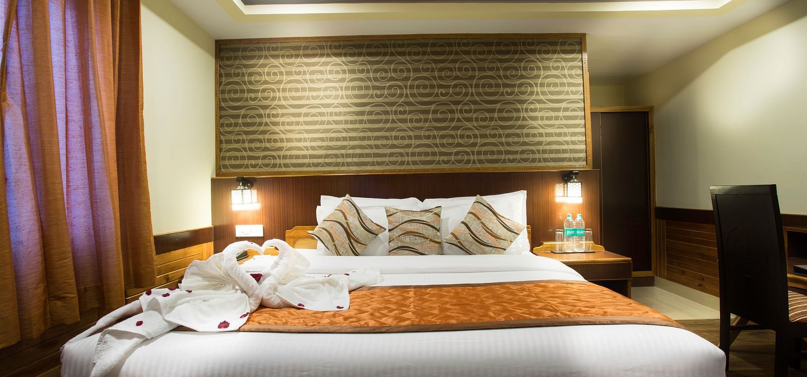 A room at New Orchid Lords Inn Gangtok contains a bed with pillows and towel swans, bedside tables with lamps and water, curtains, and a desk with a chair.