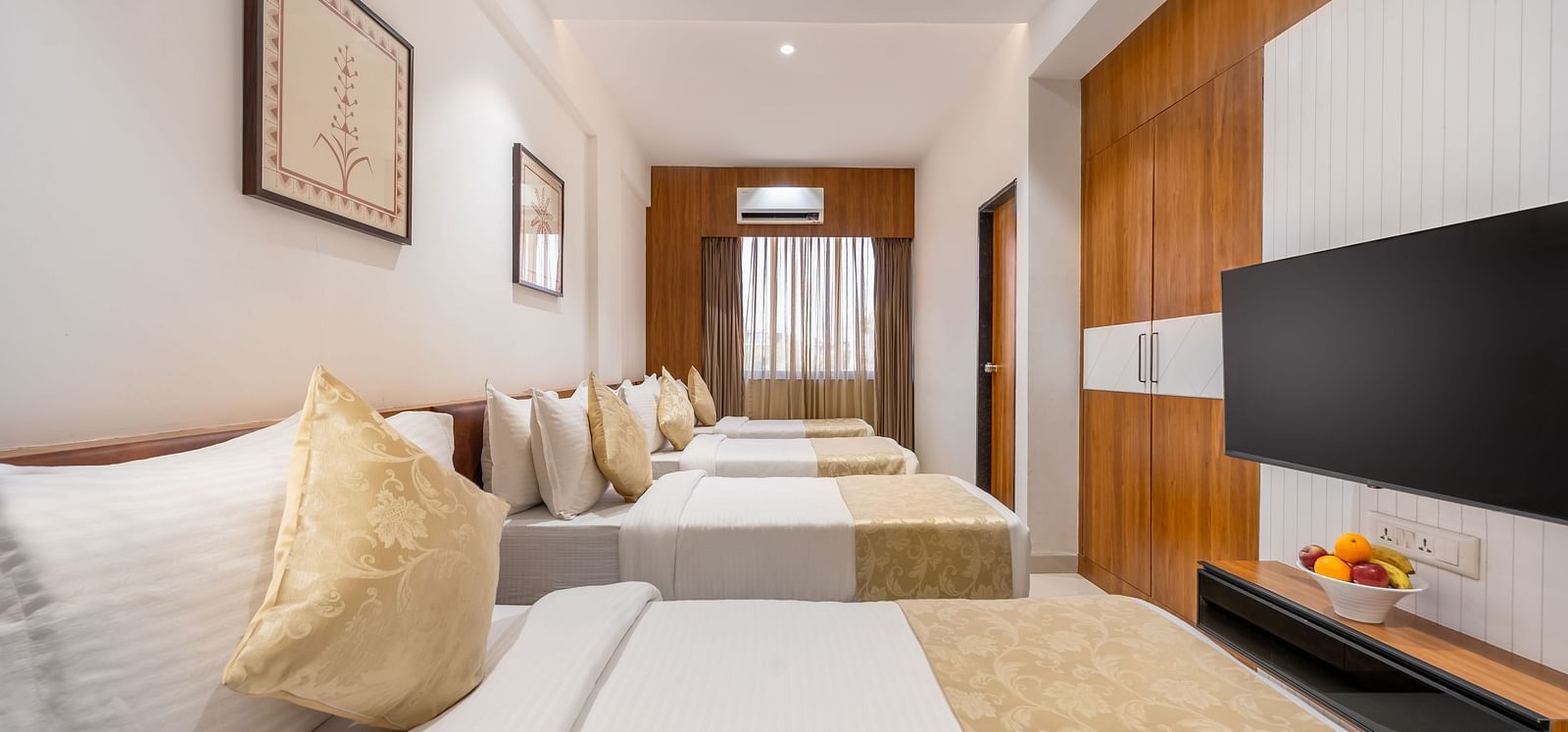 A hotel room contains four neatly made single beds, a mounted television, and simple framed artwork on the walls - Lords Eco Inn Ambaji.