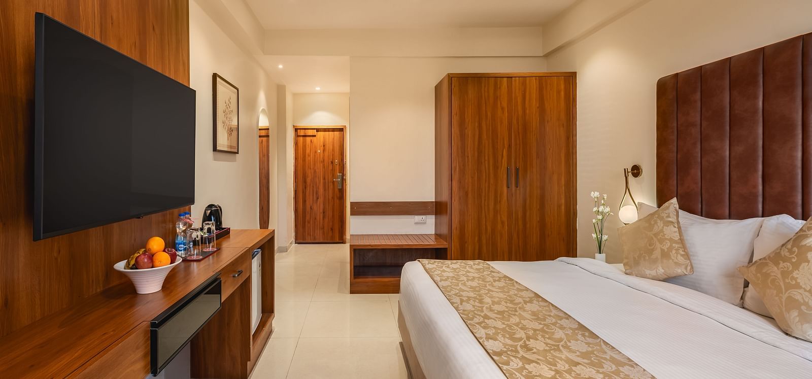 A well-appointed hotel room features a comfortable king-size bed, a mounted television above a desk with fruit, and a wooden wardrobe - Lords Eco Inn Ambaji.