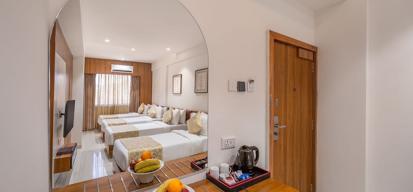 A hotel room is partially visible through a large mirror above a wooden dresser with fruit and a kettle - Lords Eco Inn Ambaji.