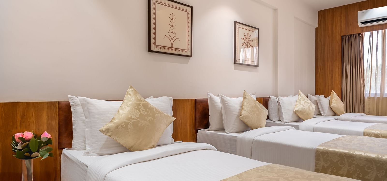 Three single beds with white linens and decorative pillows line a hotel room with framed artwork and a window - Lords Eco Inn Ambaji.