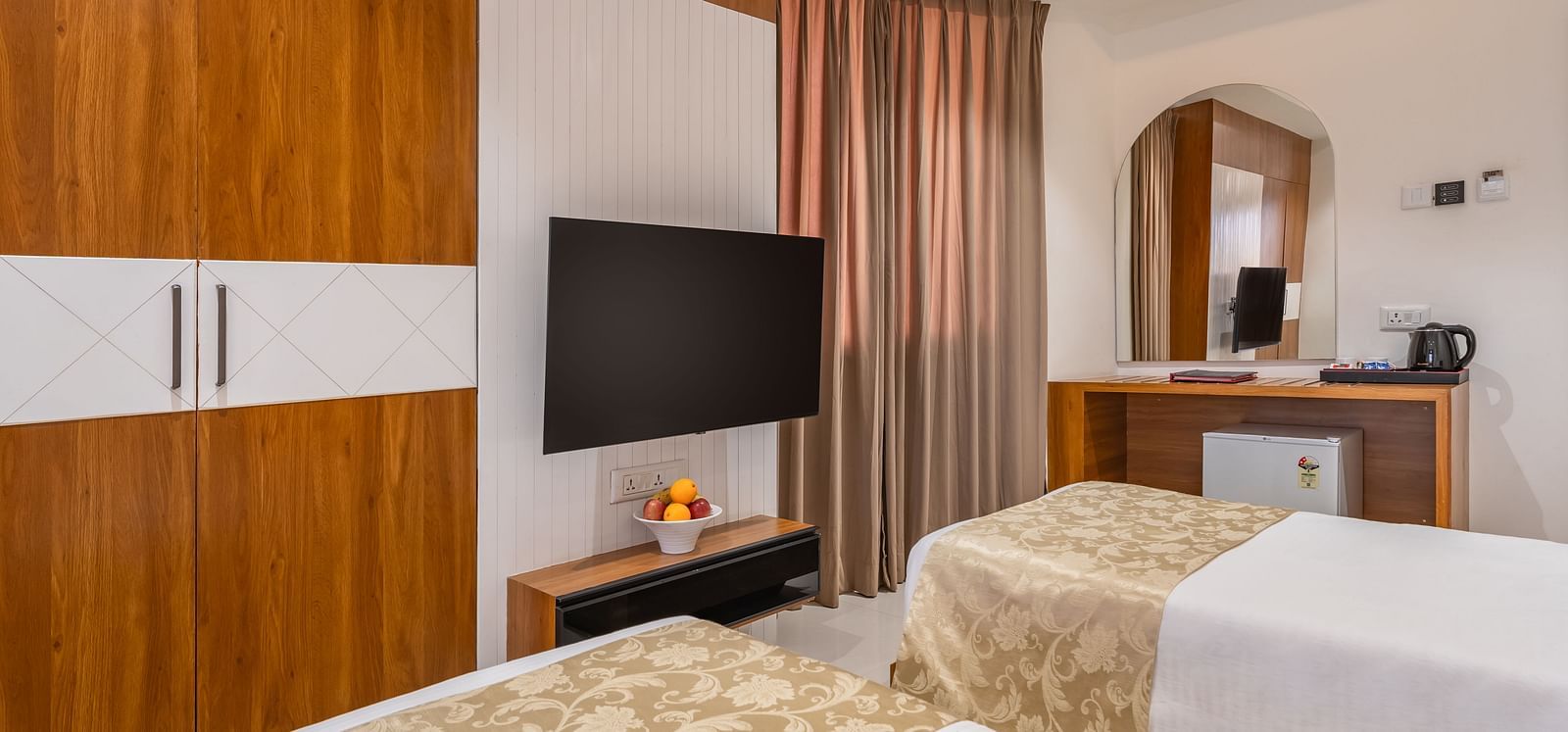 A hotel room features two single beds, a wall-mounted television, a wardrobe, and a desk with a mirror - Lords Eco Inn Ambaji.