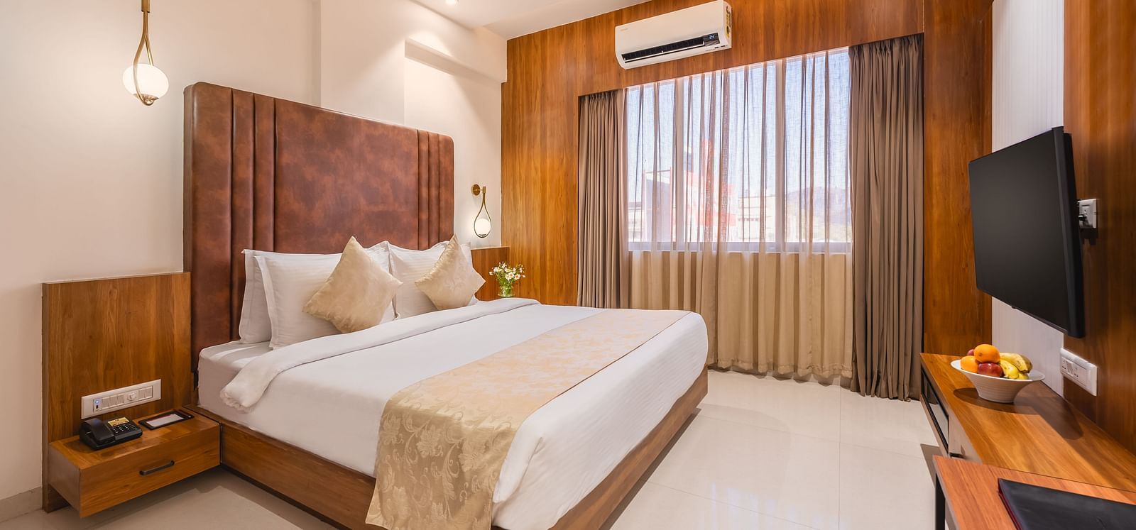 A comfortable king-size bed with soft pillows and a decorative throw faces a mounted television in a well-lit hotel room with a window - Lords Eco Inn Ambaji.
