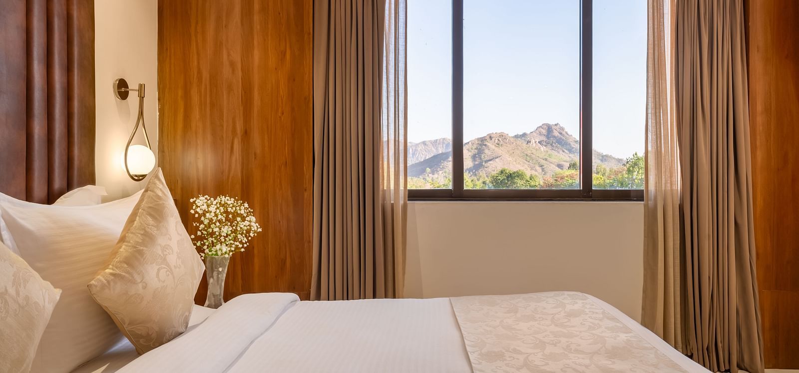A comfortable king-size bed with soft pillows and a throw is positioned in front of a large window revealing a scenic mountain view - Lords Eco Inn Ambaji.