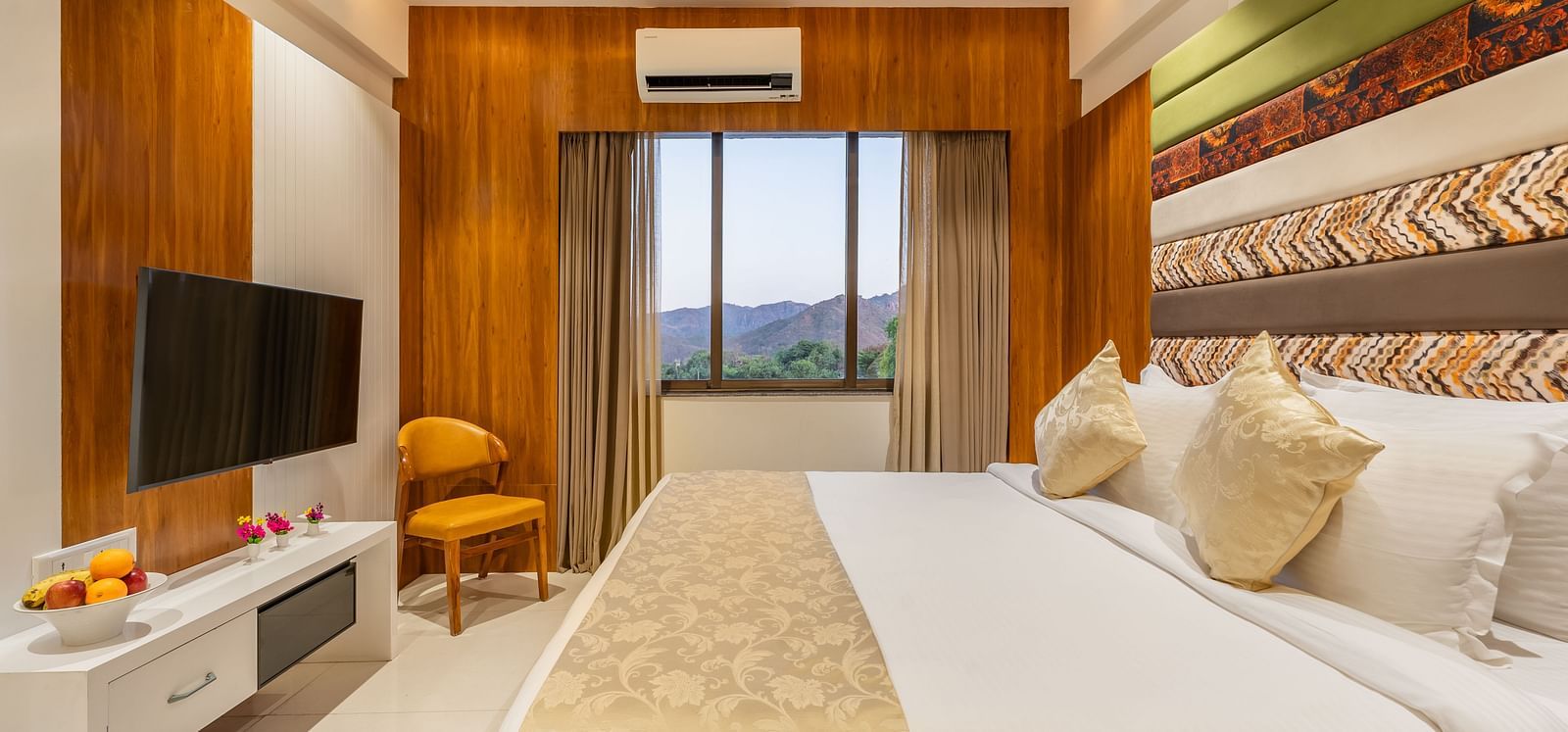 A well-appointed hotel room features a king-size bed, a mounted television, a cushioned chair, and a view through the window - Lords Eco Inn Ambaji.