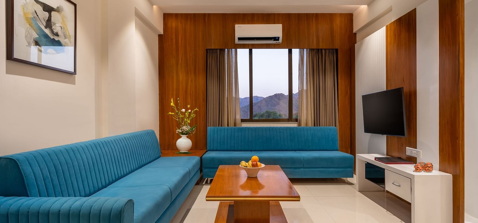 A living area features two sofas, a wooden coffee table with fruit, a television, and framed artwork - Lords Eco Inn Ambaji.