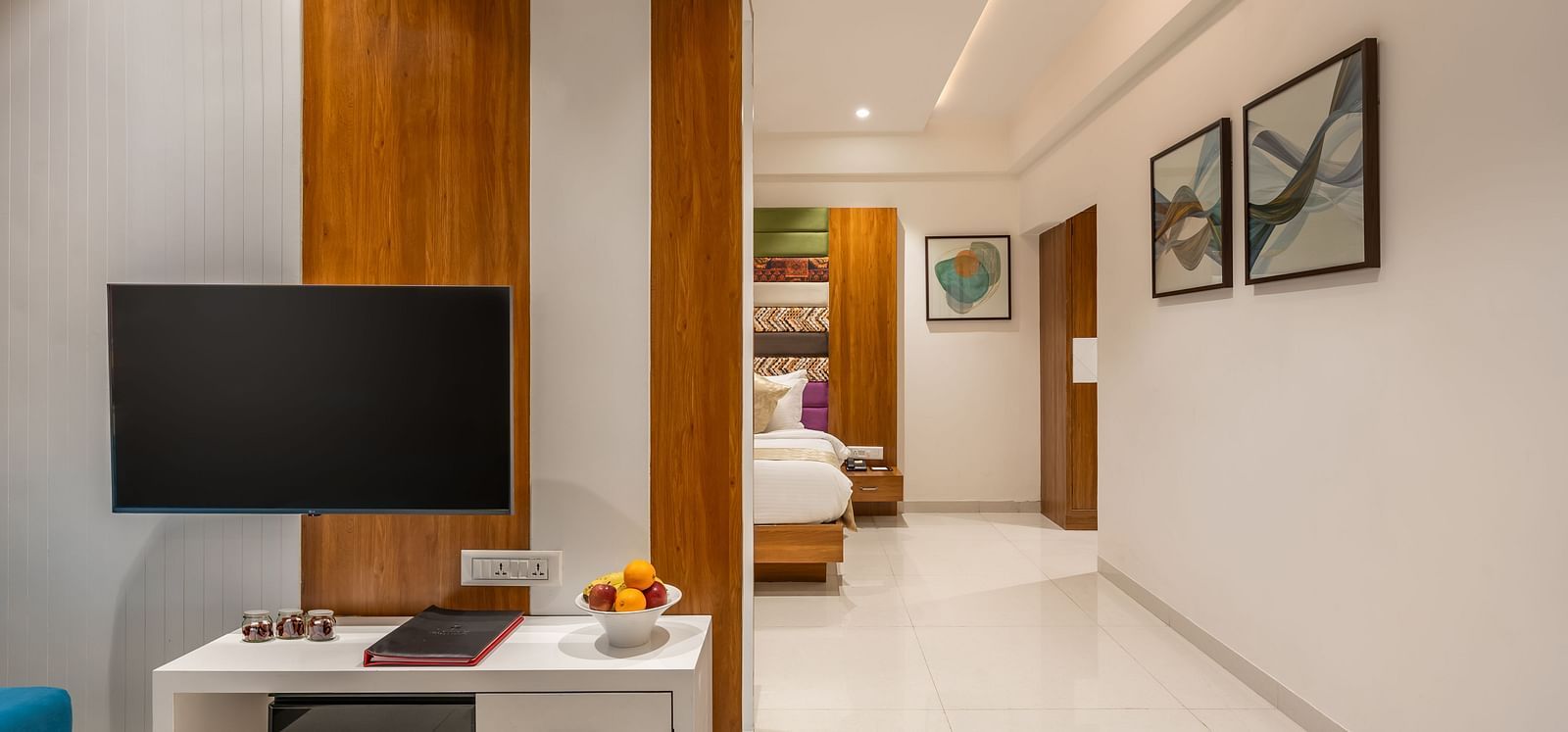 A hotel suite shows a mounted television above a desk with fruit, leading to a bedroom area and a hallway with artwork - Lords Eco Inn Ambaji.