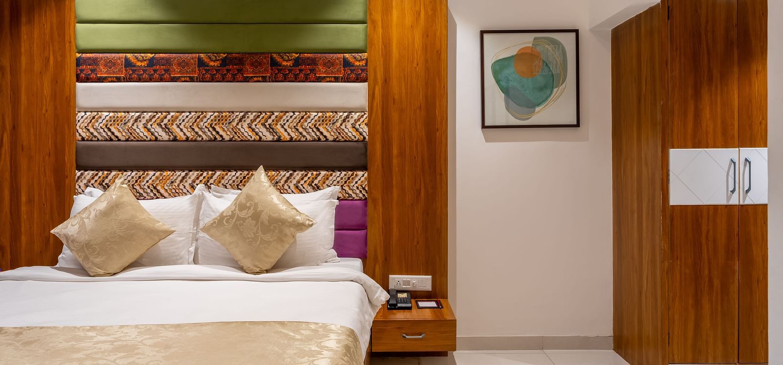 A comfortable bed with decorative pillows and a throw is positioned next to a wooden nightstand and wardrobe - Lords Eco Inn Ambaji.