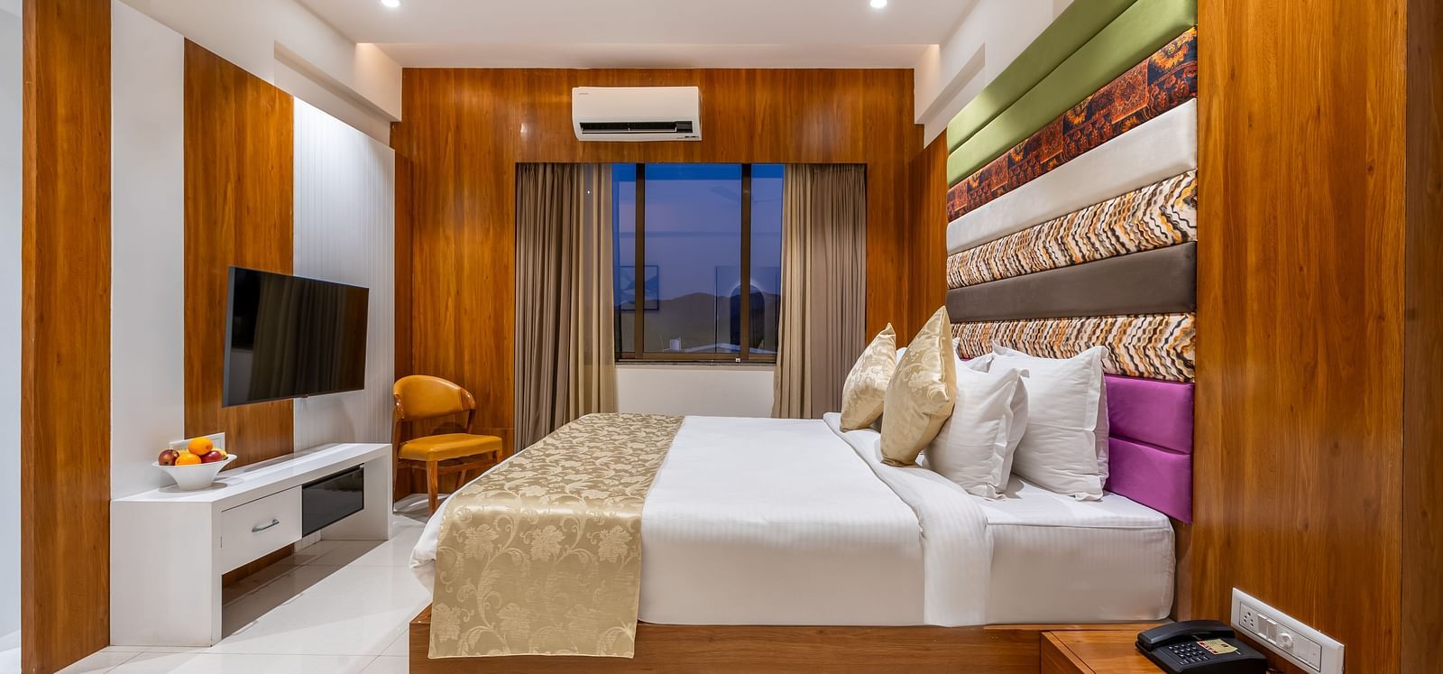 A hotel room showcases a large bed with decorative pillows and throws, a mounted TV, and a window with curtains - Lords Eco Inn Ambaji.