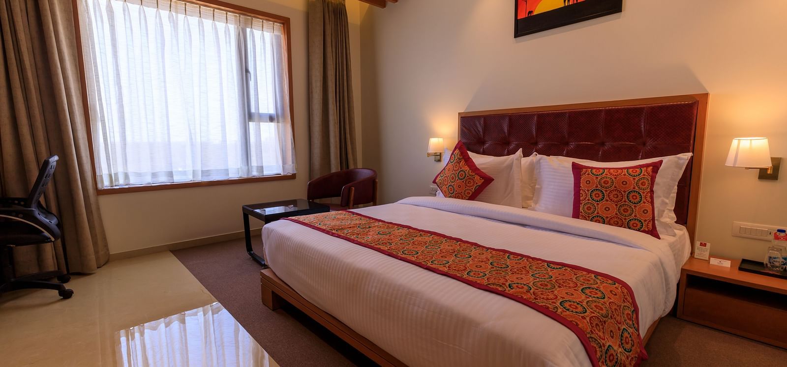 A well-lit hotel room at Lords Eco Inn Apple Gate features a double bed with patterned accents, a desk, a chair, and window curtains.