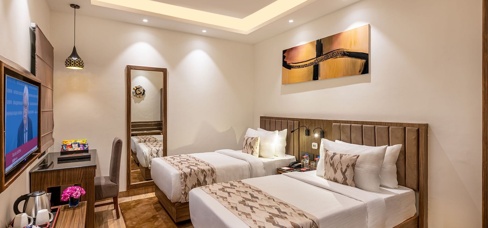 A hotel room with two twin beds, a flat-screen TV, and a work desk. It features a unique tray ceiling with lighting, a ceiling fan, and colourful abstract artwork - Lords Eco Inn Dehradun.
