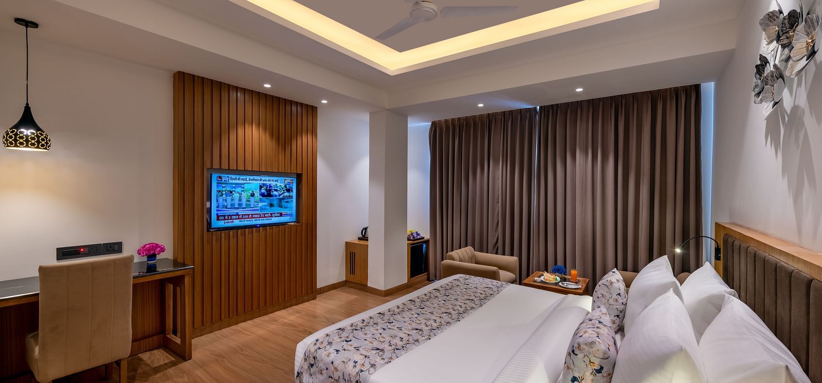 A spacious hotel room featuring a bed, a work desk with a chair, a wall-mounted TV, a ceiling fan, and large windows with heavy drapes - Lords Eco Inn Dehradun.