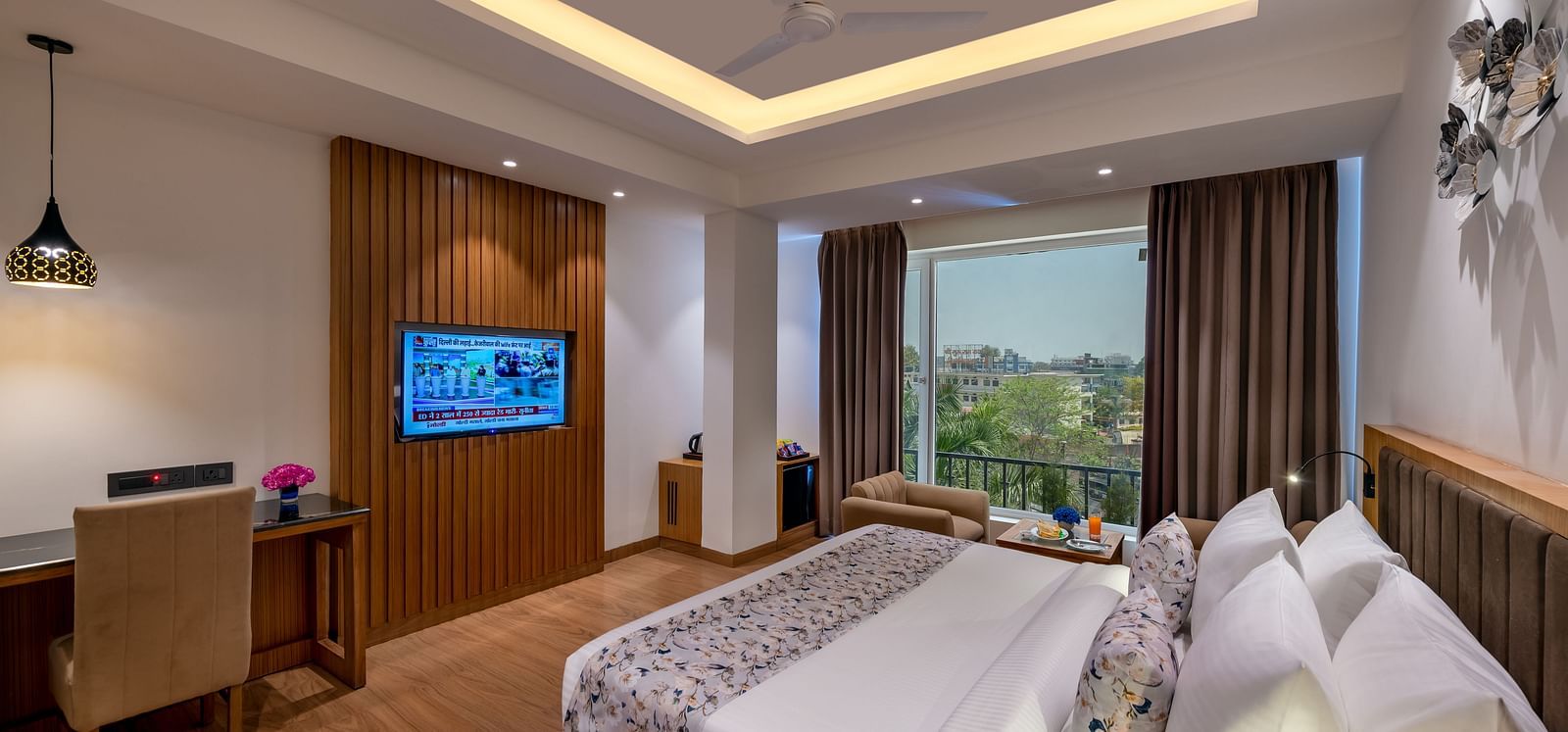 Hotel room with a king bed, desk, and TV, featuring a large window with a city view, dark drapes, and a modern tray ceiling with lighting - Lords Eco Inn Dehradun.