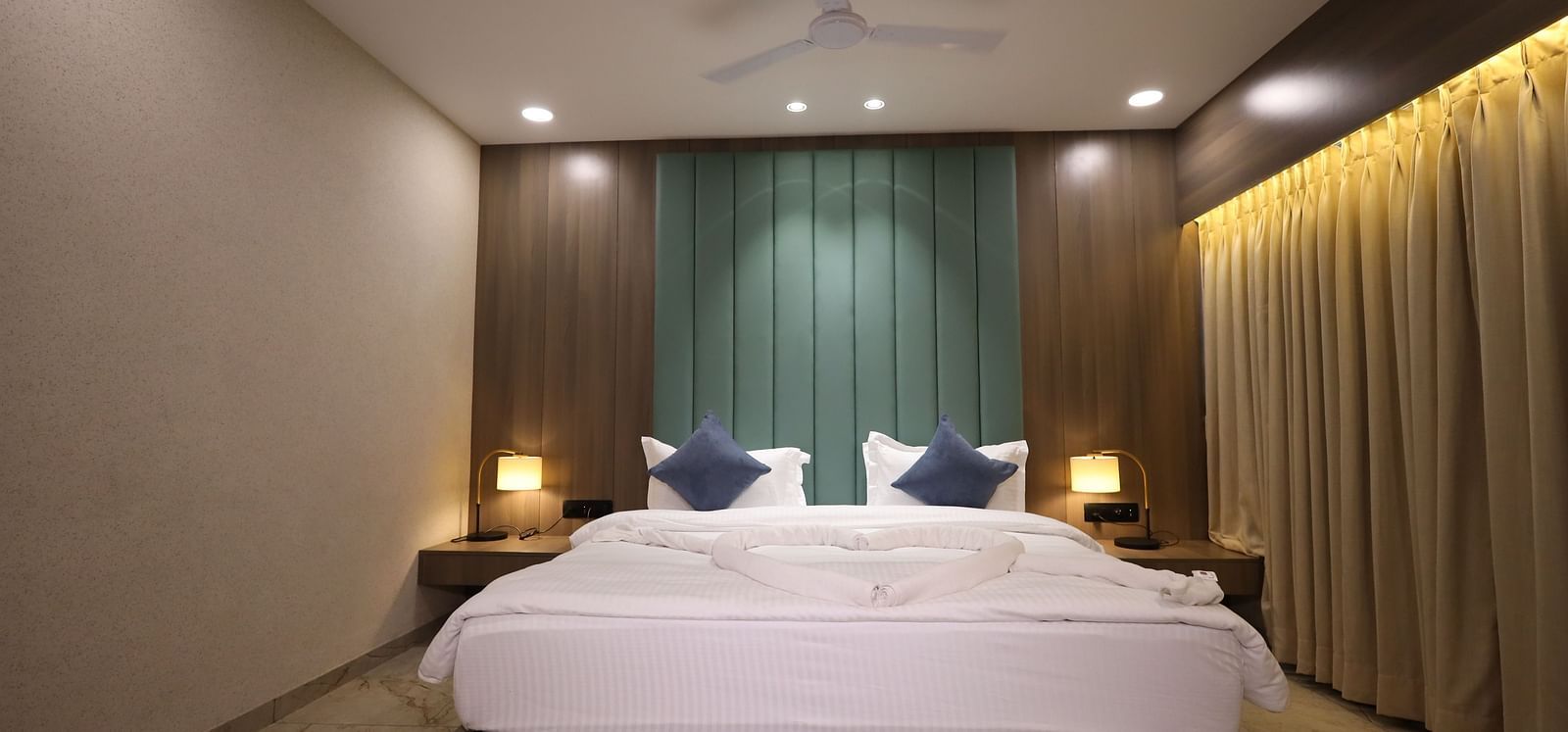 A well furnished room with a king size bed, blue pillows, bedside tables with lamps and expansive beige curtains at Lords Eco Inn Navsari.