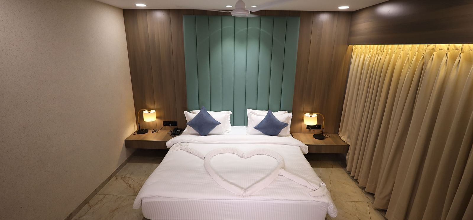 A cosy room with a heart shaped decorated bed featuring bedside tables with lamps, blue pillows and beige curtains at Lords Eco Inn Navsari.