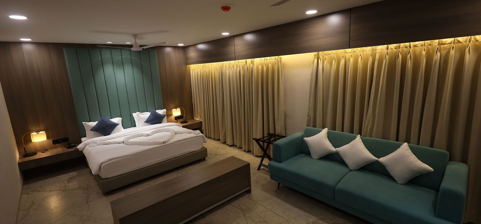 A cosy suite with a king size bed, blue sofa with cushions, bedside tables with lamps, and curtains expanding across the back of the sofa to the bed at Lords Eco Inn Navsari.