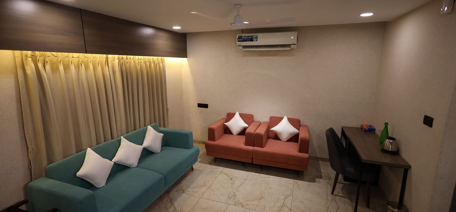 A cosy living space with two blue sofa with cushions set against draped curtains, red armchairs and a small table with a chair and amenities at Lords Eco Inn Navsari.