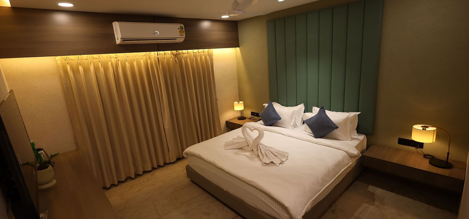 Modern designed room with a king size bed against the patterned accent wall decorated with swan-shaped towels, bedside tables and lamps, illuminated curtains and an elongated desk with potted plants and a flat screen T.V. at Lords Eco Inn Navsari.