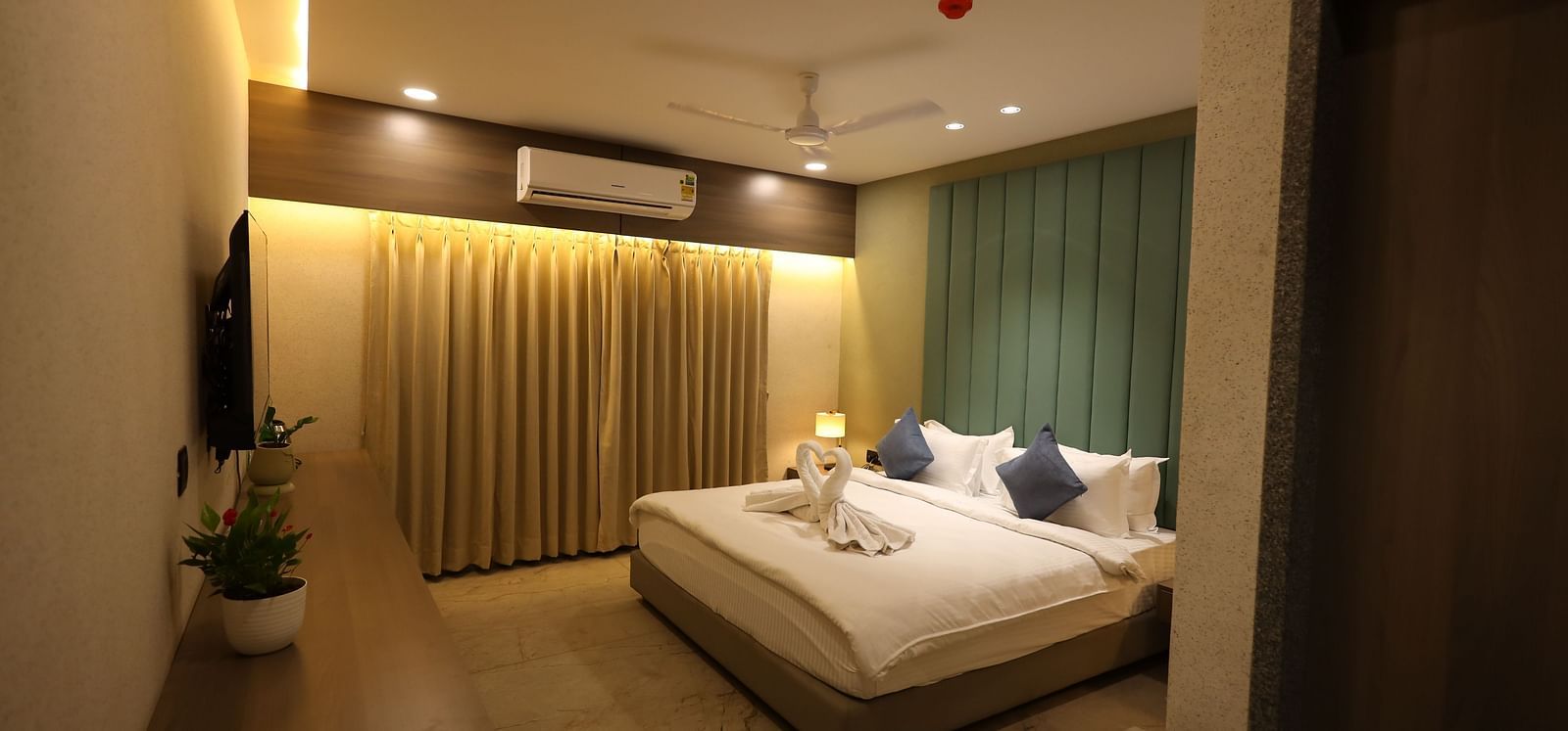 A hotel room in Navsari features a bed with folded towels, bedside lamps, and a long dresser with plants - Lords Eco Inn Navsari.