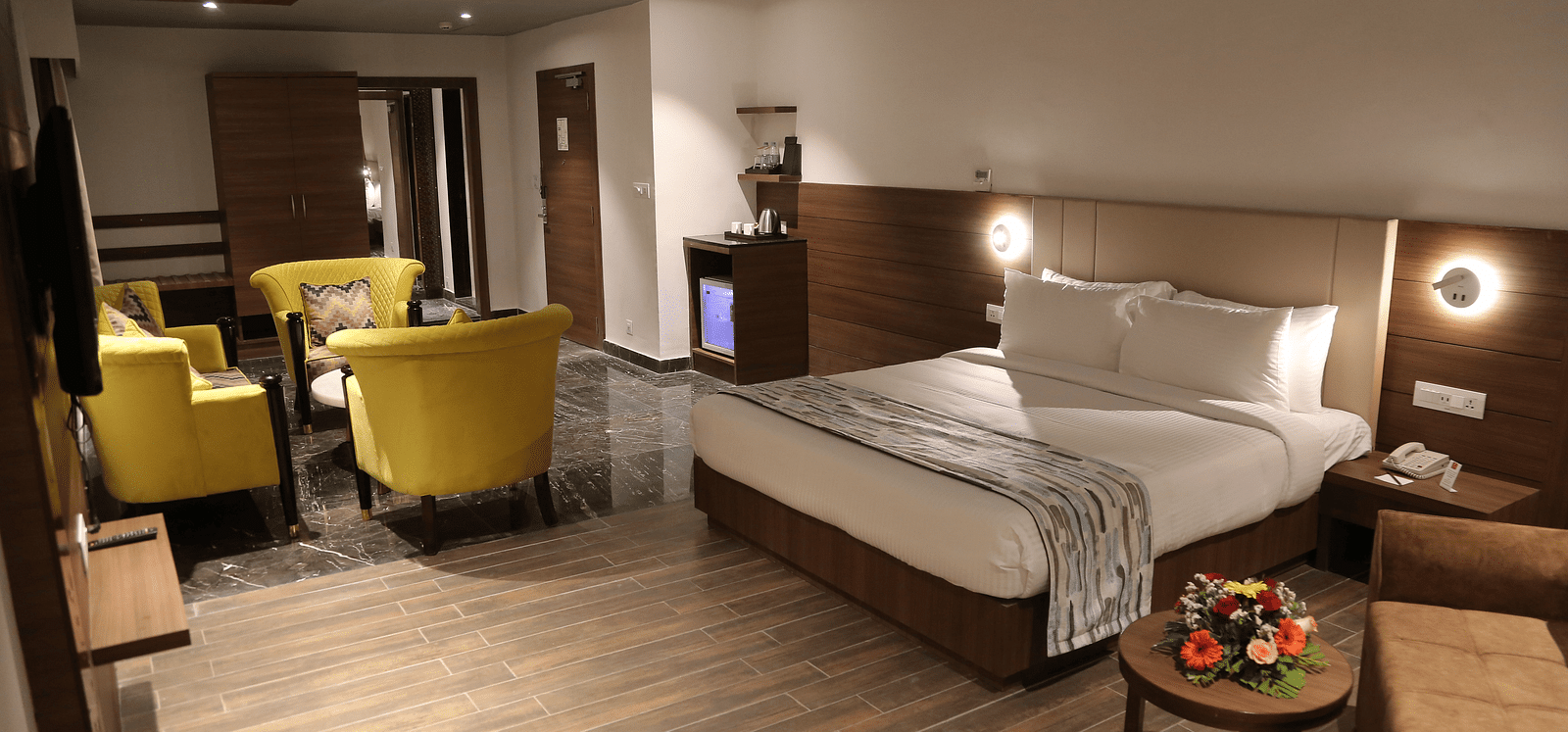 A spacious hotel room features a neatly made bed, a seating area with yellow armchairs, and modern decor under soft lighting.