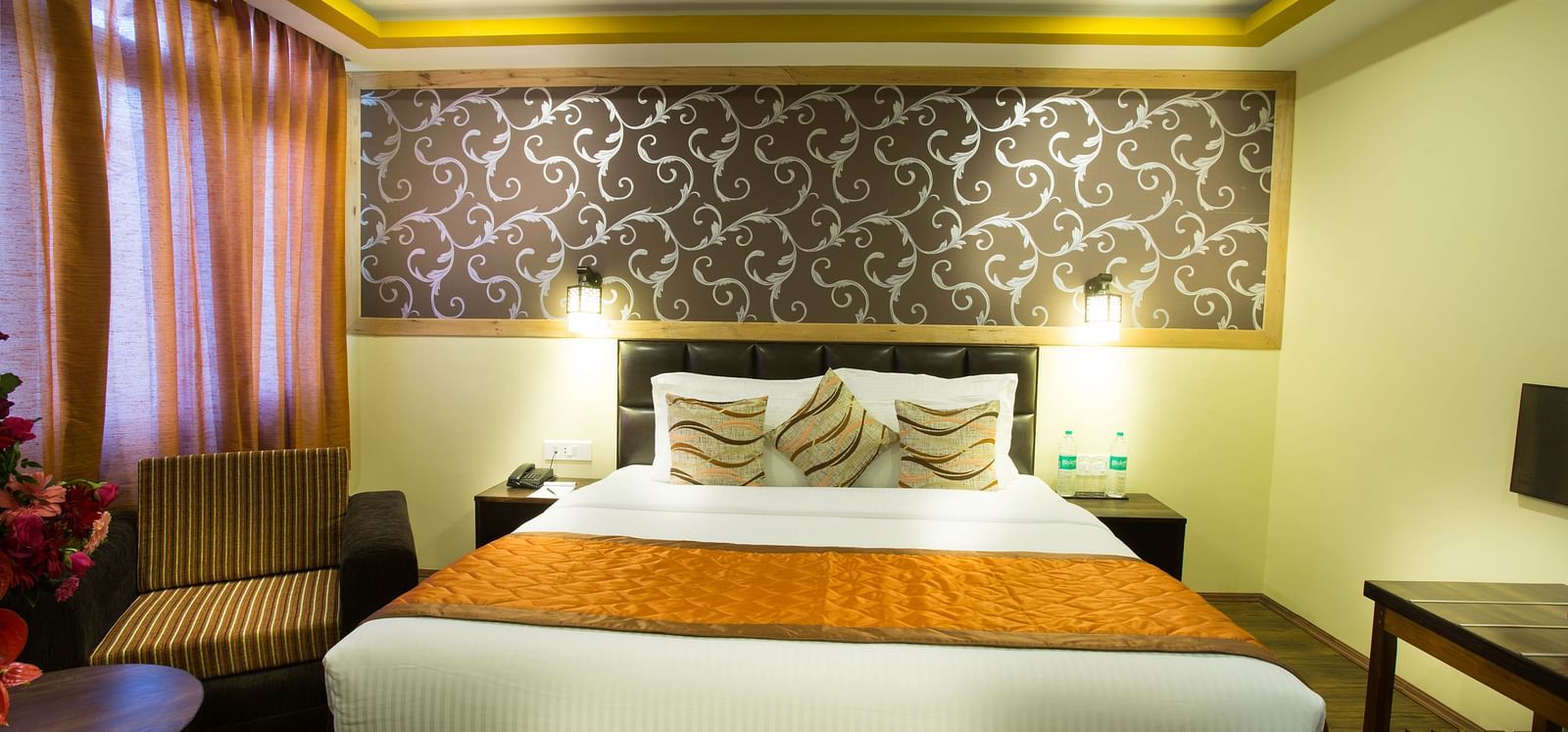 A room at New Orchid Lords Inn Gangtok contains a bed with pillows, bedside tables with lamps and phones, curtains, an armchair, and a coffee table with flowers.