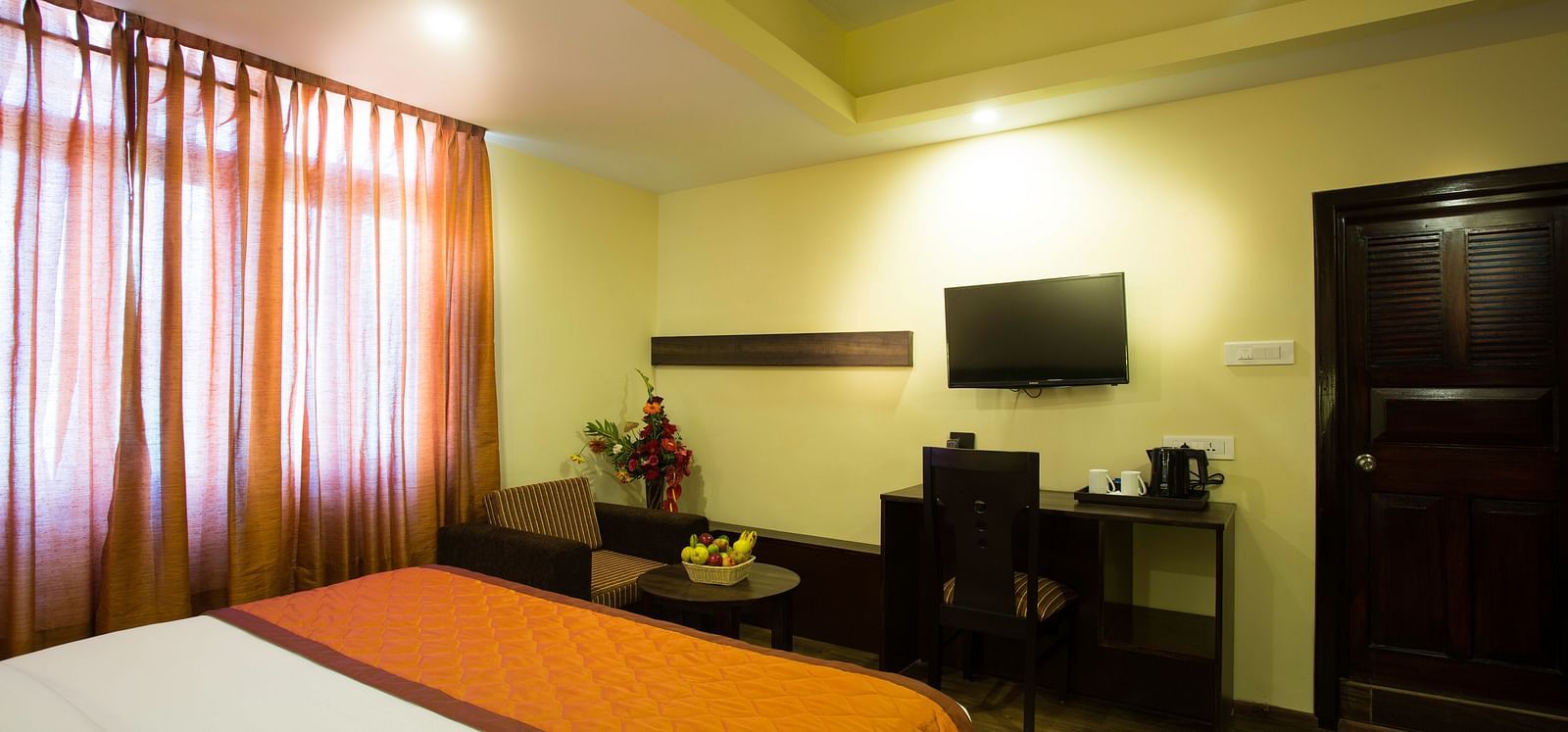 The image shows a bed, curtains, a desk, a chair, a television, a door, and some decorative items in a room -  New Orchid Lords Inn Gangtok.