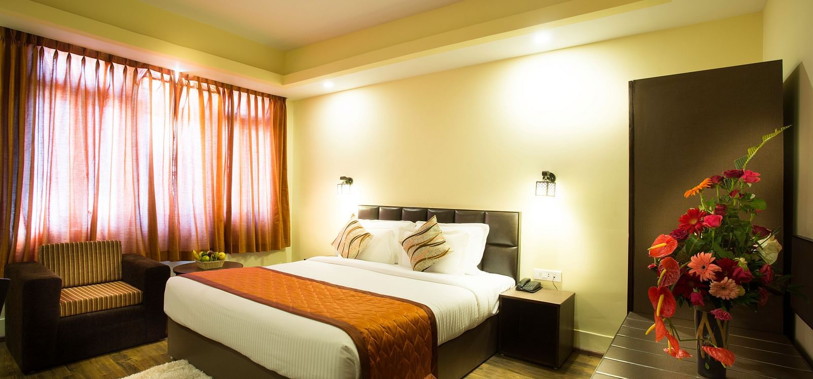 A room at New Orchid Lords Inn Gangtok contains a bed with pillows, a bedside table with a lamp and phone, curtains, an armchair, a rug, and a vase of flowers.