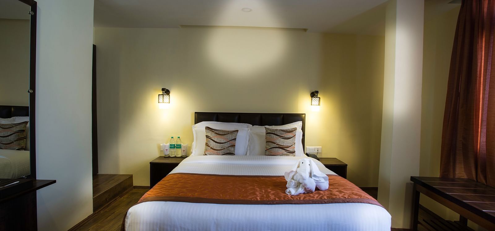 A room at New Orchid Lords Inn Gangtok contains a bed with pillows and towel swans, bedside tables with lamps and water, curtains, and a desk.