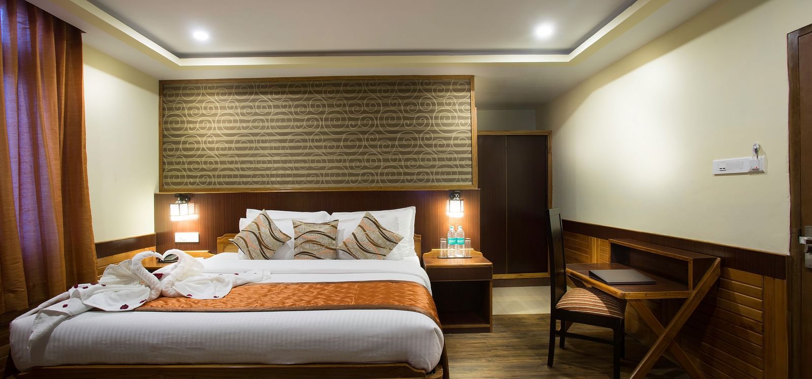 A room at New Orchid Lords Inn Gangtok contains a bed with pillows and towel swans, bedside tables with lamps, curtains, and a desk with a chair.