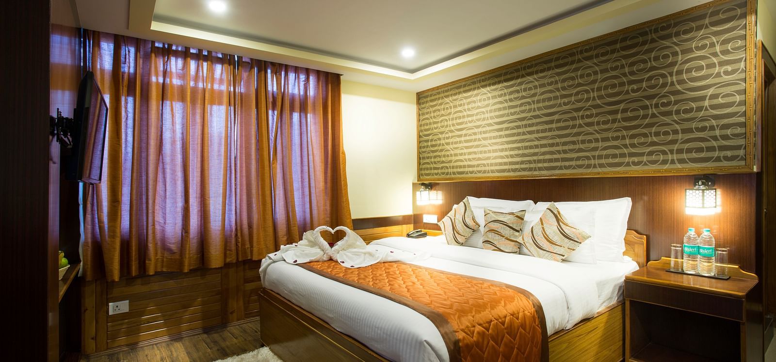 A room at New Orchid Lords Inn Gangtok contains a bed with pillows and folded towels, bedside tables with lamps and water bottles, curtains, and a rug.        A room at New Orchid Lords Inn Gangtok contains a bed with pillows and decorative towel swans, be