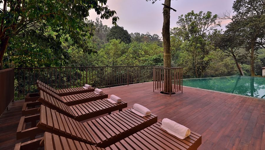 An outdoor seating area features wooden chairs with trees and a building in the background. - Abad Brookside Lakkidi, Wayanad