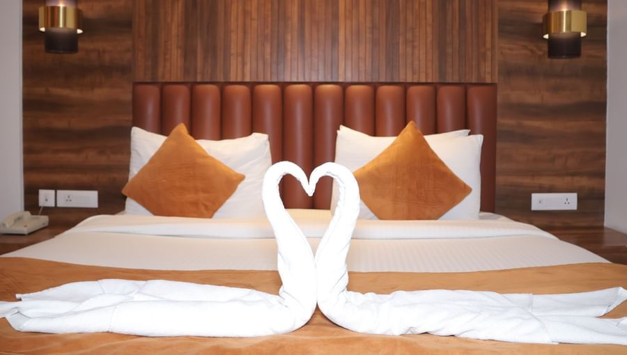 A pair of towel swans at the centre of the bed with pillows, cushions, and a wooden headboard at Best Western, Vrindavan.