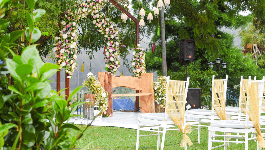 A charming outdoor wedding or event area featuring a rustic wooden archway adorned with white and pink flowers, with white chairs and tables set on a grassy area, surrounded by lush green plants.