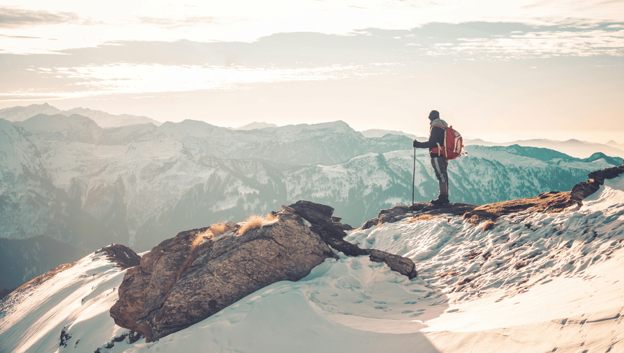 A lone hiker with a backpack standing on a snowy mountain ridge, looking out over a vast mountain range at sunrise.