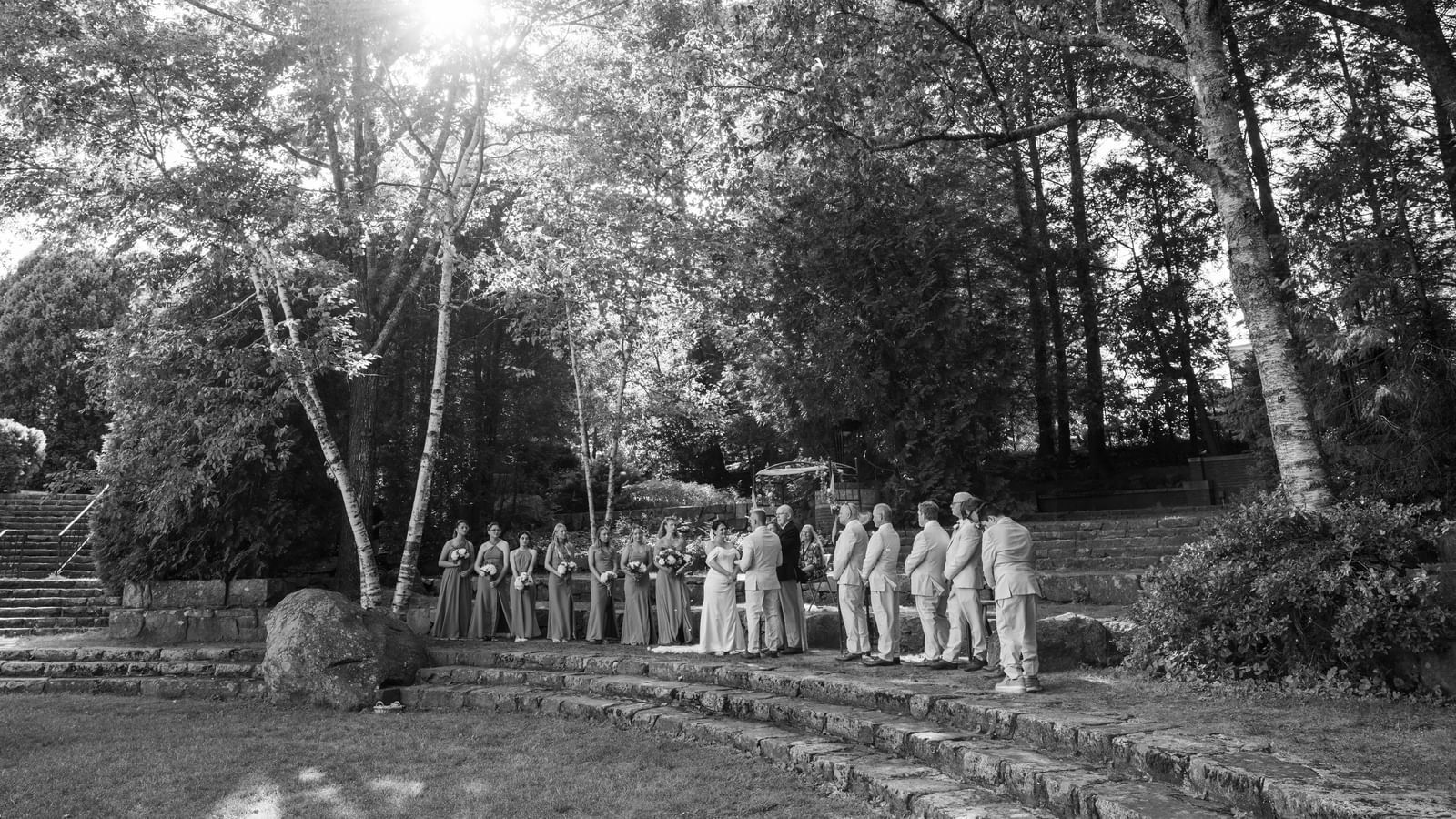 Wedding ceremony at Hartstone Inn with the couple, officiant, bridesmaids, and groomsmen standing on stone steps surrounded by trees and sunlight filtering through.