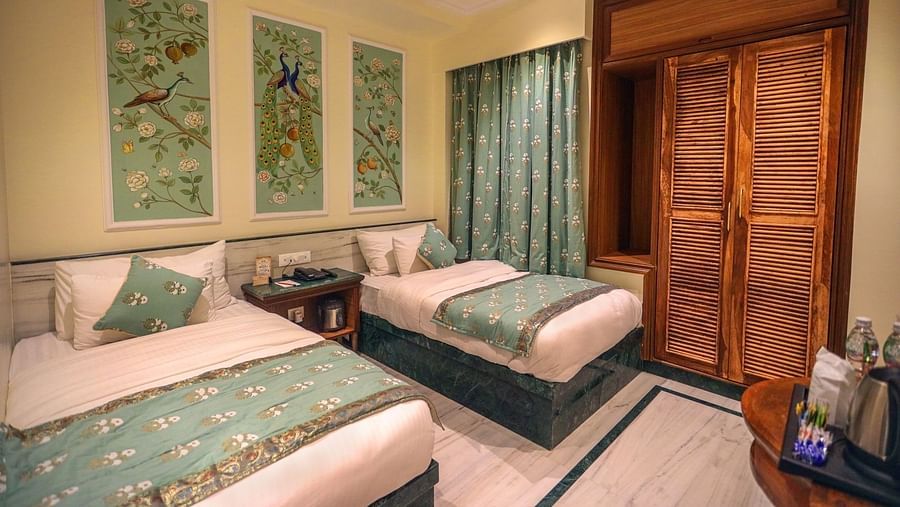 Diagonal view of the spacious room with twin beds placed side by side, a wooden bedside table in the middle, three photo frames above the headboard, and wooden storage | Panna Vilas Udaipur