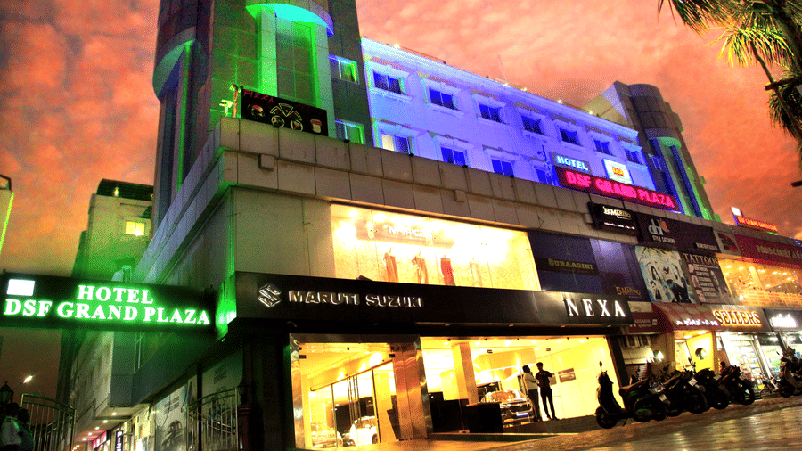 The illuminated exterior of DSF Grand Plaza, Tuticorin at night with coloured facade lighting, hotel signage, and a lit showroom entrance below, framed by a palm tree.