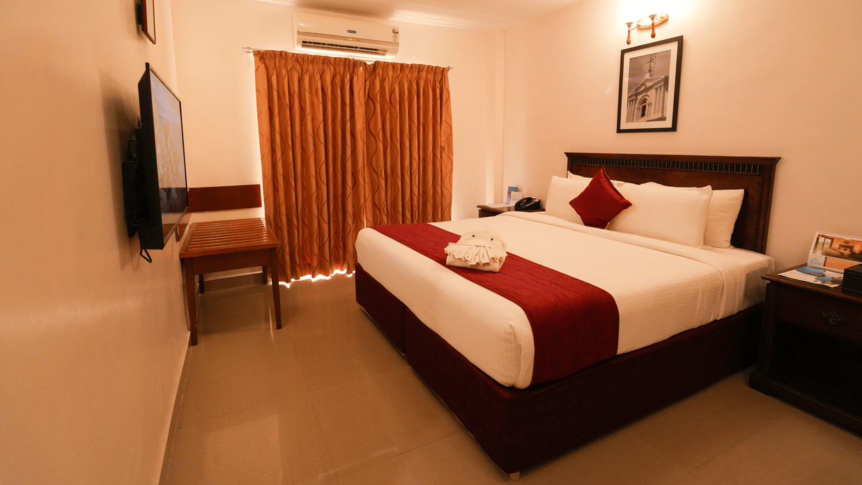 MGM Vailankanni Residency - Deluxe Room - Queen Bed. Image of the well-made bed, bedside tables, and floor-to-ceiling curtains.